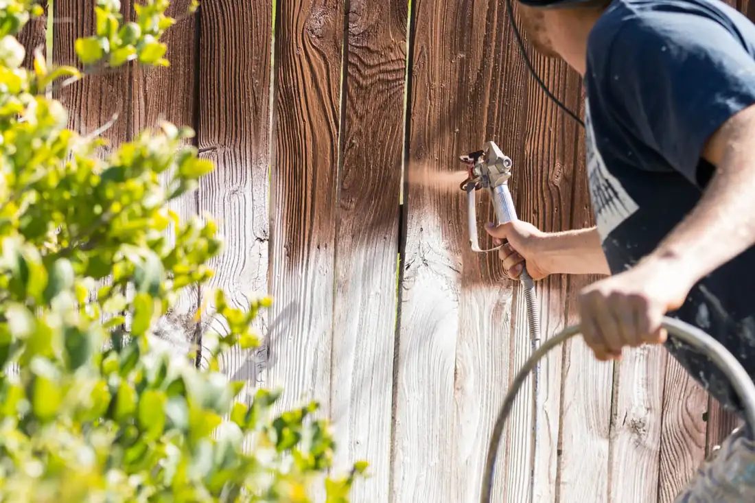 Worker spraying stain onto a weathered wood fence
